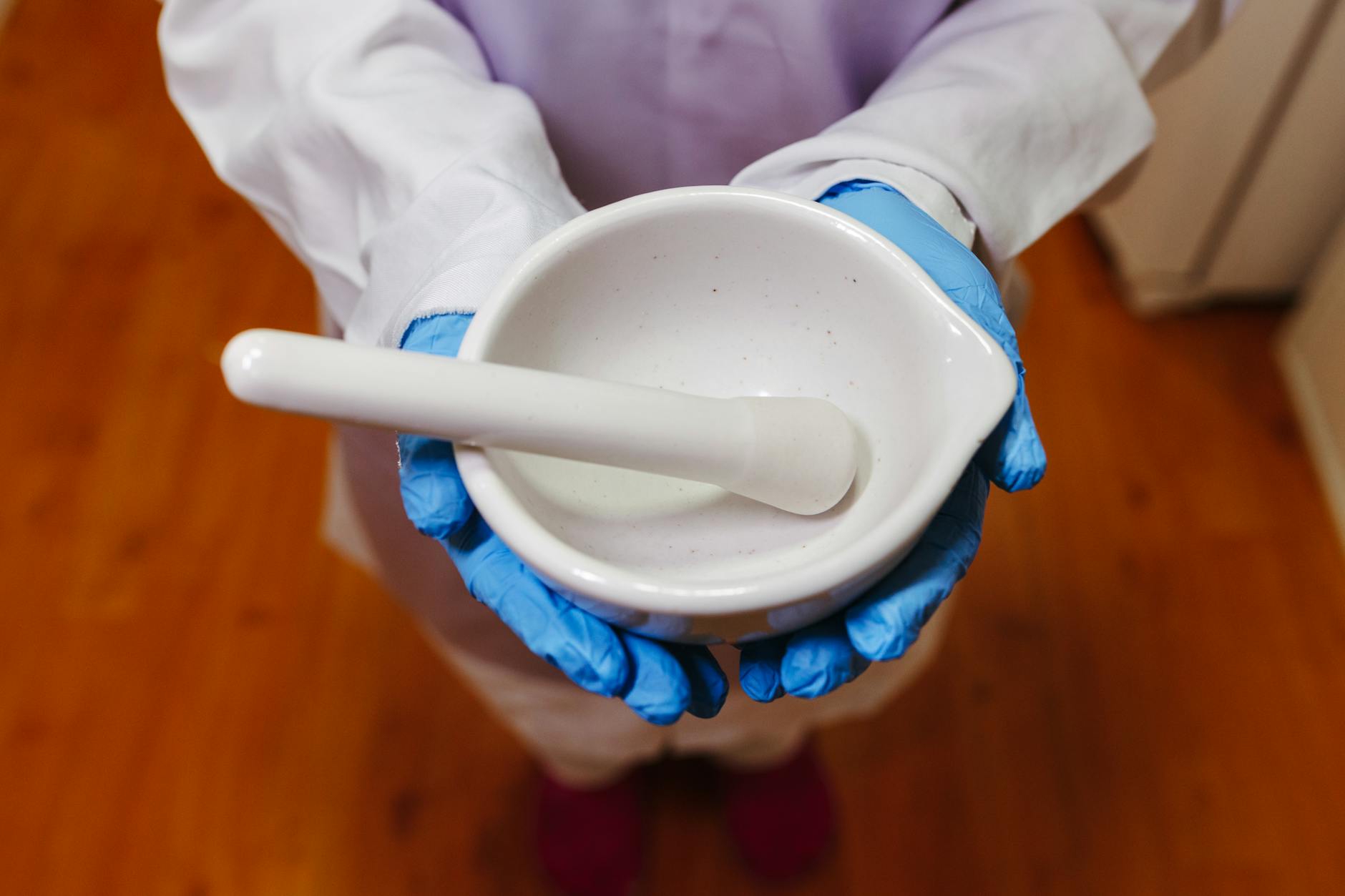Pharmacist holding a white mortar and pestle, wearing blue gloves and lab coat indoors
